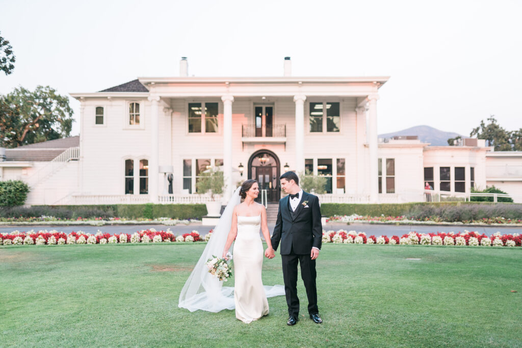 A bride and groom stand holding hands on a lawn in front of a large white mansion, with flowerbeds in the background.