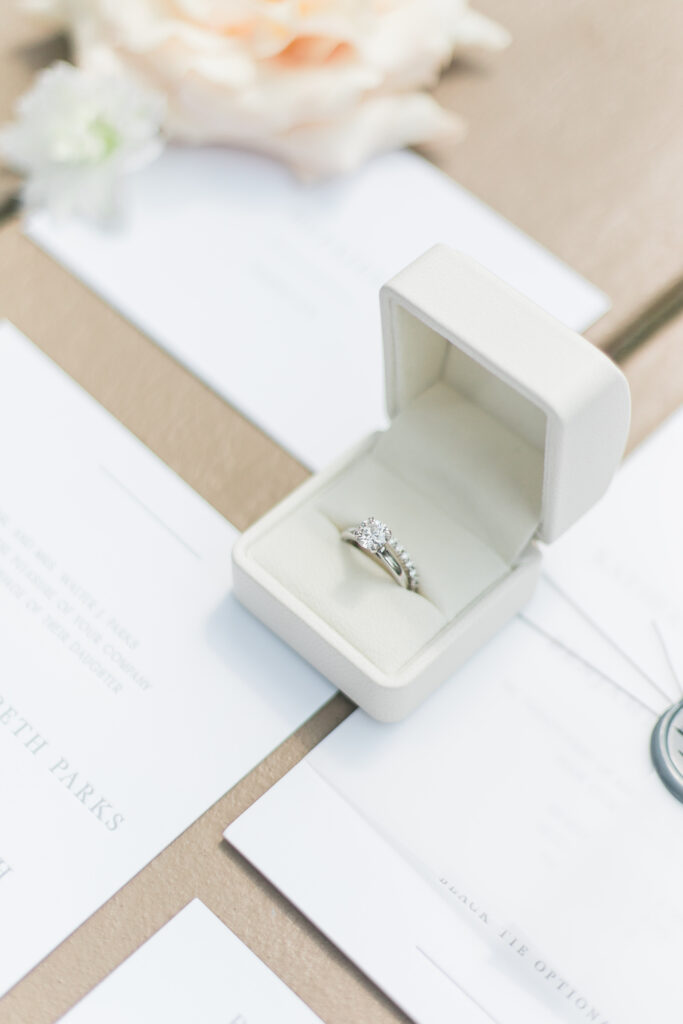 A diamond engagement ring and wedding band in a white box, placed on a table with wedding invitations and a blush-colored flower.