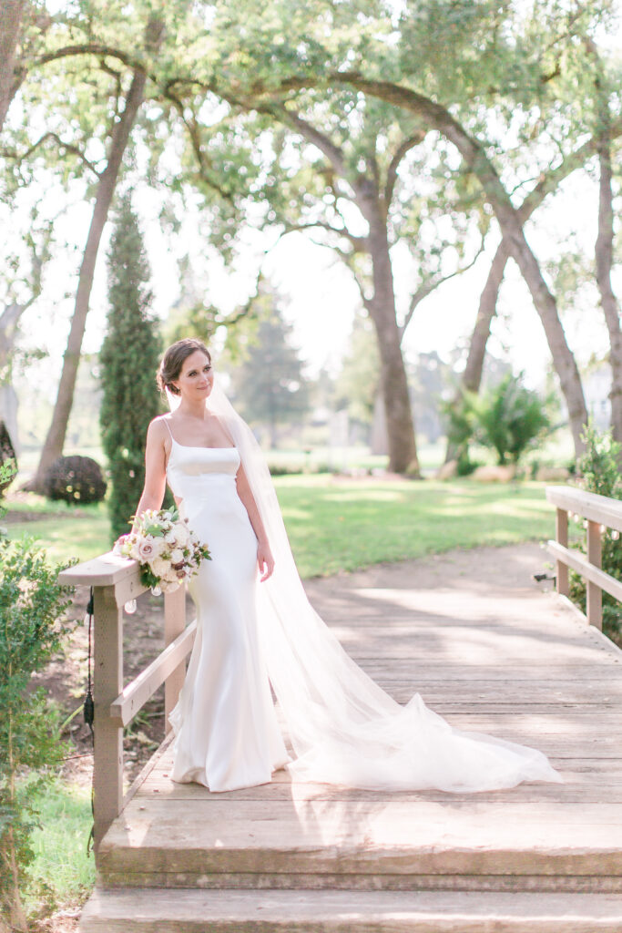 A bride in a white gown with a long veil stands on a wooden bridge outdoors, holding a bouquet, surrounded by greenery and trees.