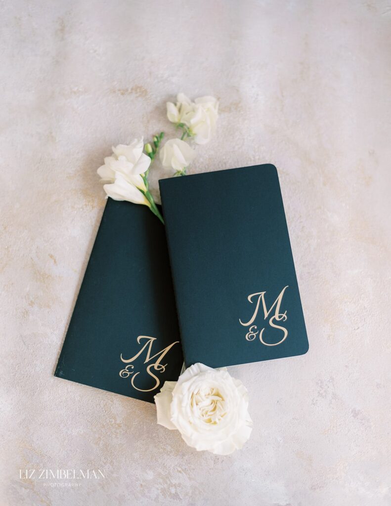 Two black notebooks with "M & S" in gold lettering are placed on a light surface, decorated with white flowers.