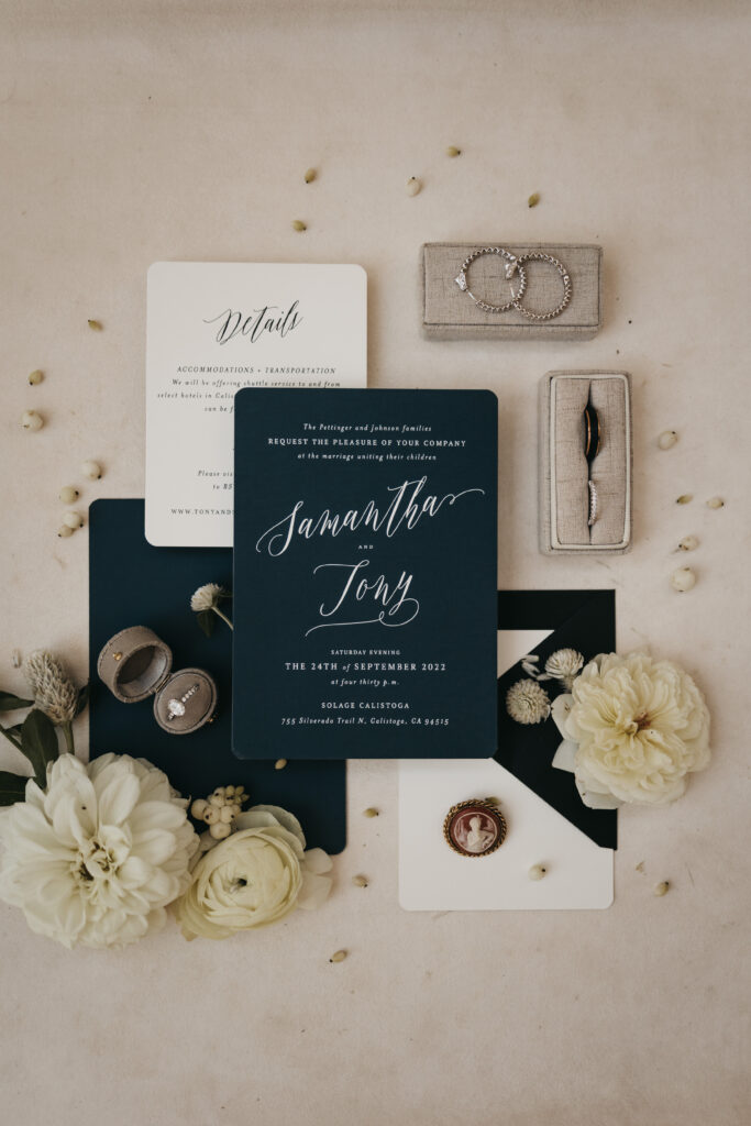 Flat lay of wedding invitation set with navy and white cards, rings, jewelry, and white flowers arranged on a light surface.