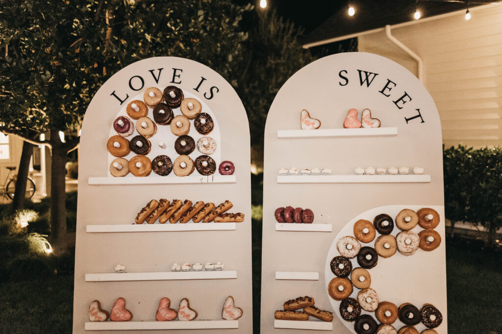 Two white boards with shelves hold an assortment of donuts and pastries arranged to spell "LOVE IS SWEET" at an outdoor event.
