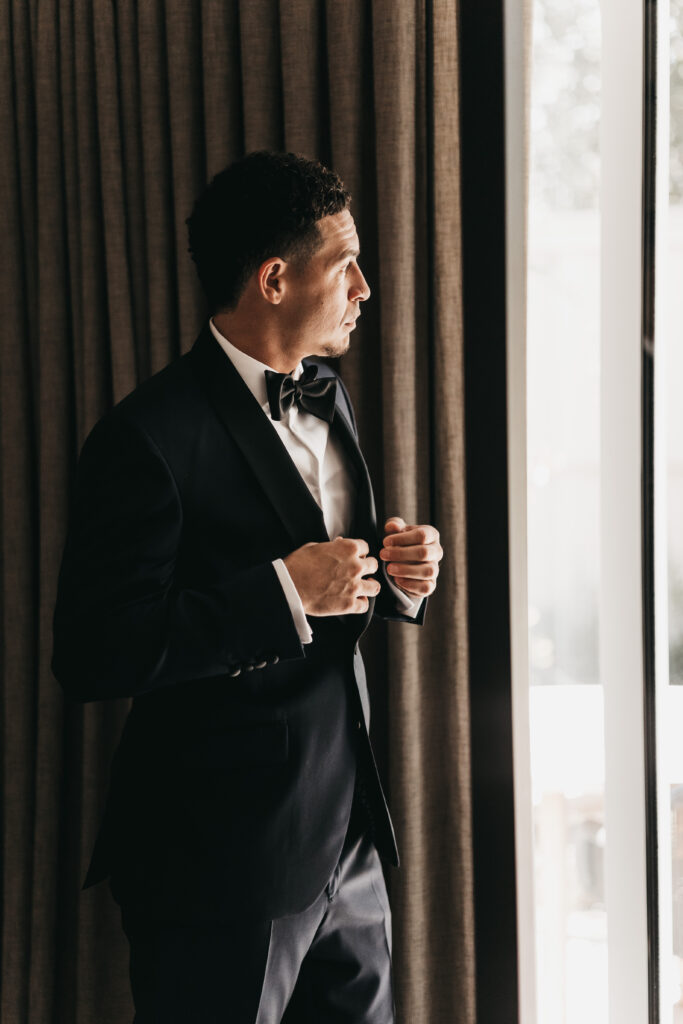 A man in a black tuxedo stands by a window, adjusting his jacket and looking outside, with curtains in the background.