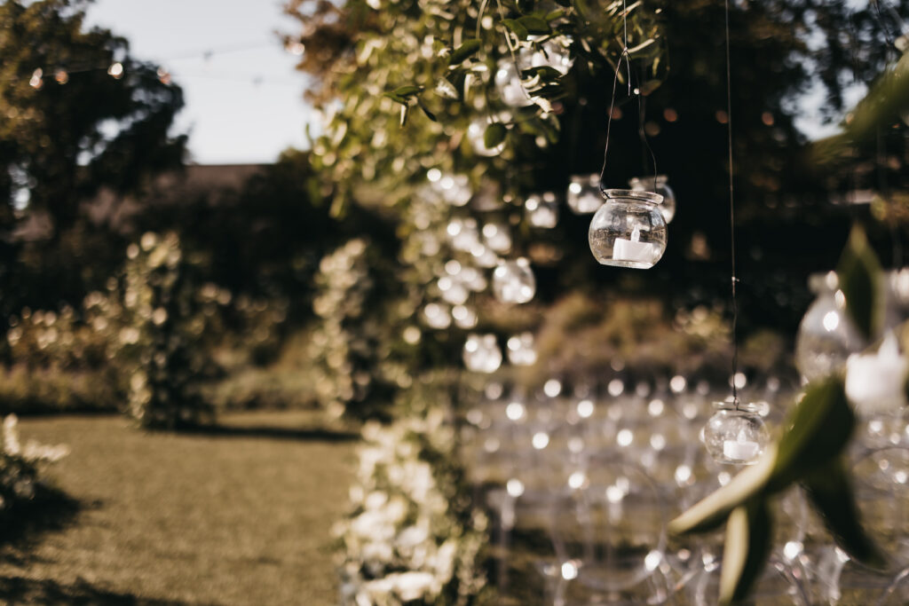Glass jars are hanging from tree branches outdoors, with sunlight filtering through greenery and rows of chairs set up in the background.
