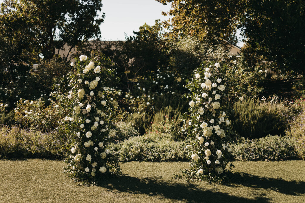 Two vertical arrangements of white flowers and greenery stand on a grassy lawn with trees and shrubs in the background.