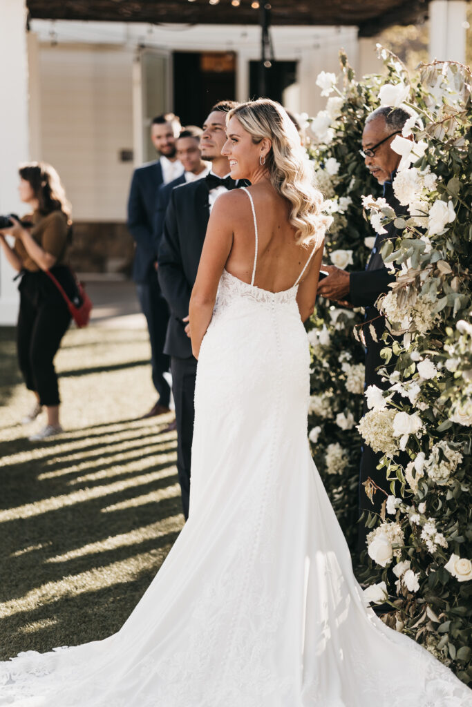 A bride in a white gown stands beside a groom during an outdoor wedding ceremony, surrounded by flowers and guests.