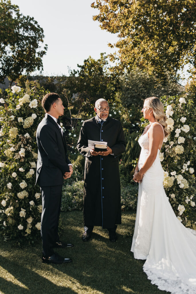 A bride and groom stand facing each other outdoors with a wedding officiant between them, surrounded by floral arrangements.