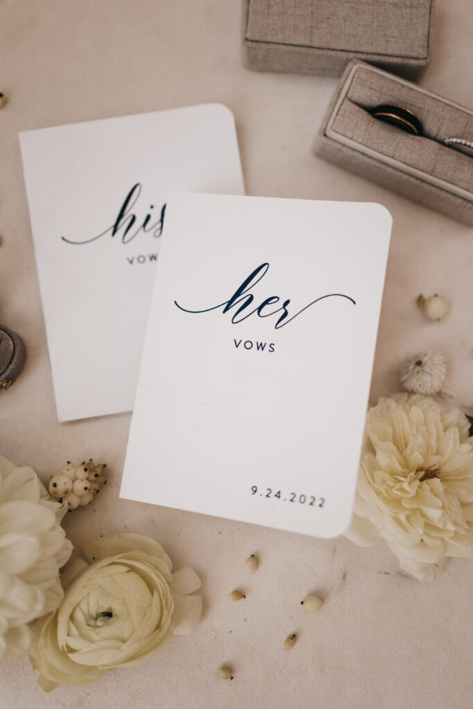 Two vow booklets labeled "his" and "her" with "9.24.2022" printed on one, surrounded by white flowers, a ring box, and a ring on a beige surface.