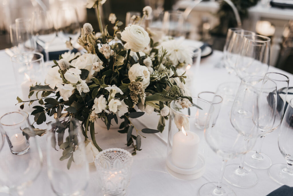 Elegant table setting with a white floral centerpiece, glassware, and lit candles on a white tablecloth.
