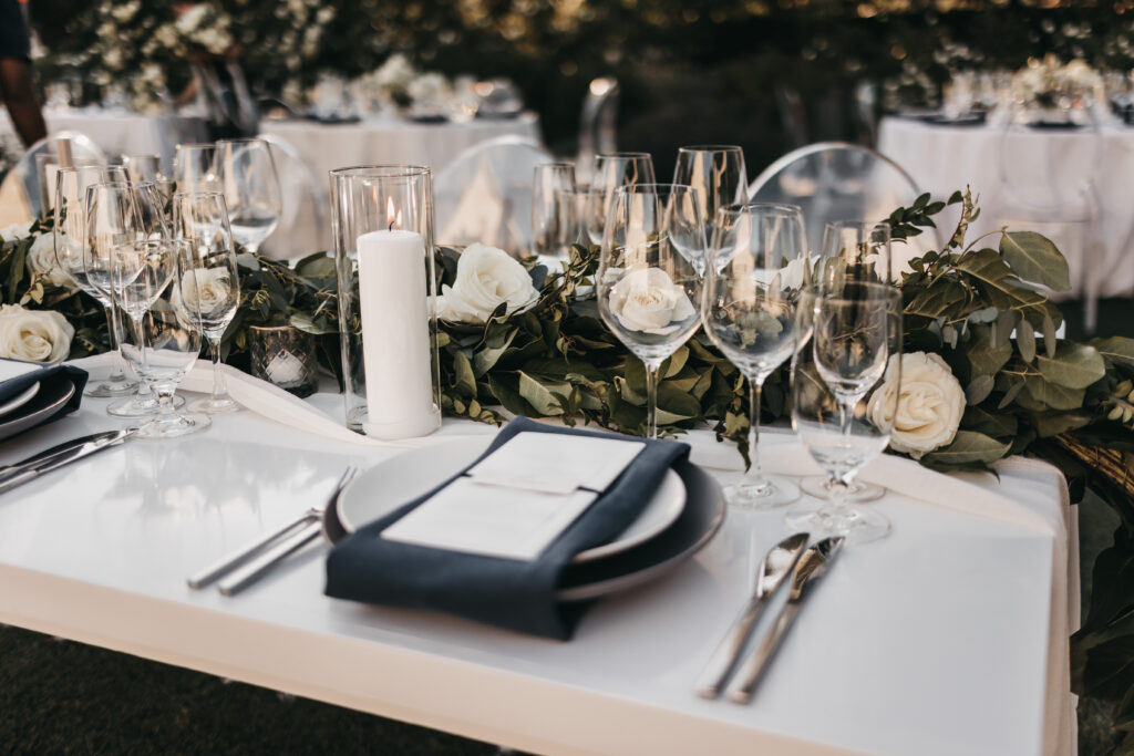 Elegant table setting with white plates, glasses, silverware, a black napkin, a lit candle, and white flowers with greenery as centerpiece.