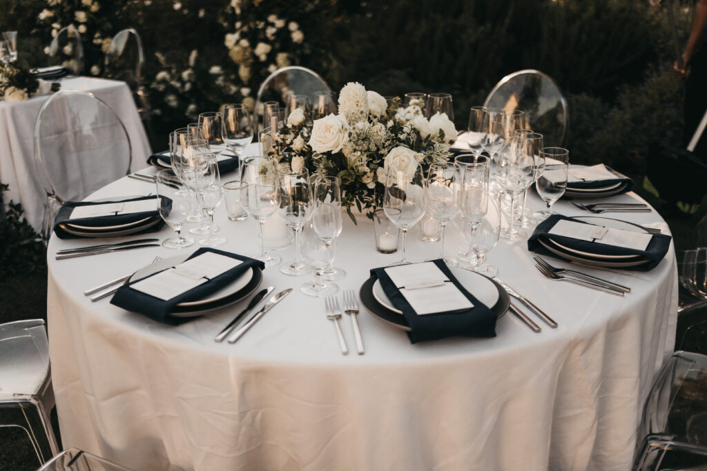 A round table set for a formal event with white tablecloth, black napkins, menus, glassware, silverware, and a floral centerpiece featuring white flowers.