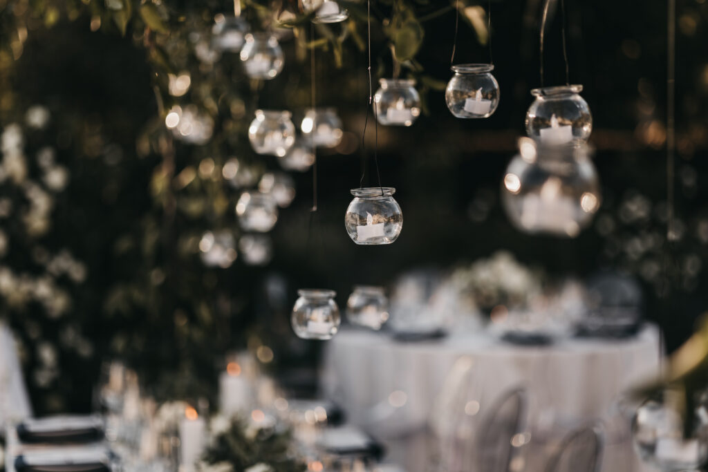 Small glass jars with candles hang from above, creating a soft, decorative ambiance at an outdoor event with blurred tables in the background.