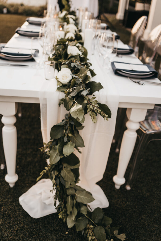 A white table set for a formal event with black napkins, glassware, and a green and white floral garland running down the center.