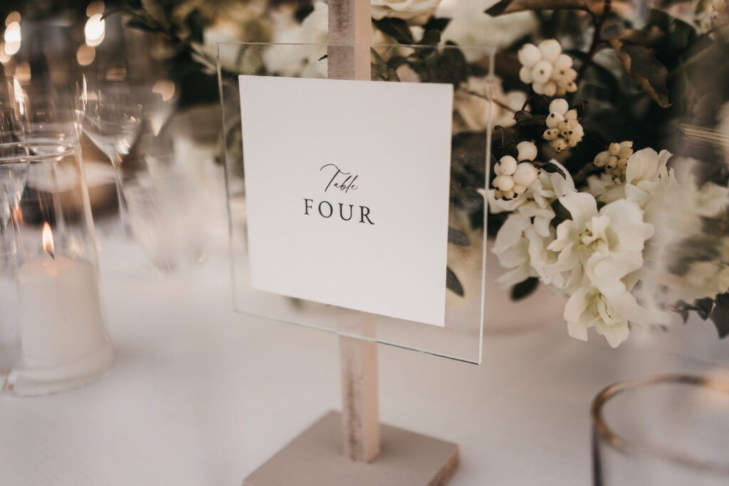 A white card with "Table Four" written on it is displayed on a holder, surrounded by white flowers and candles on a decorated table.