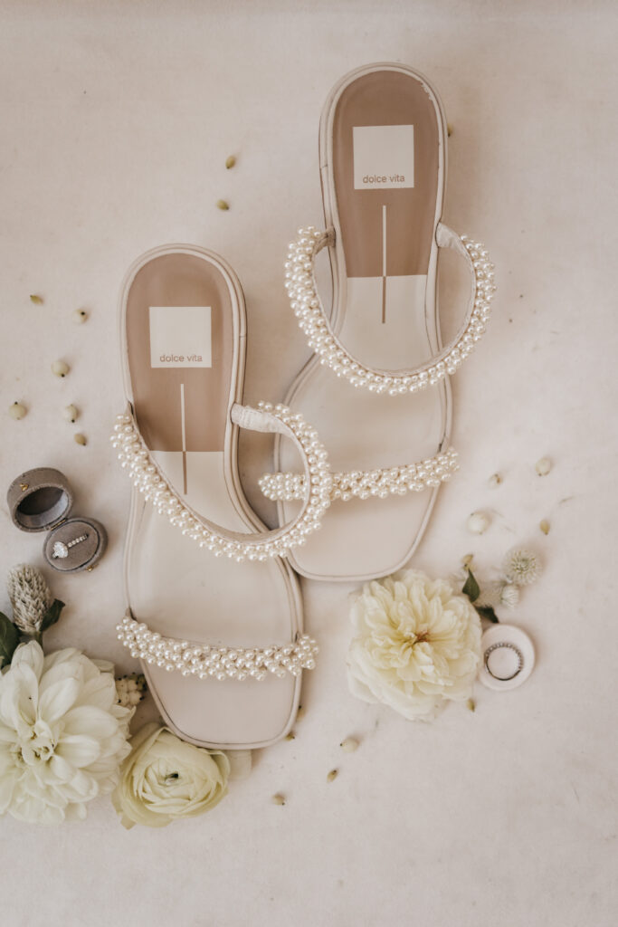 A pair of ivory sandals with pearl embellishments, surrounded by white flowers, green buds, and two ring boxes containing wedding rings.