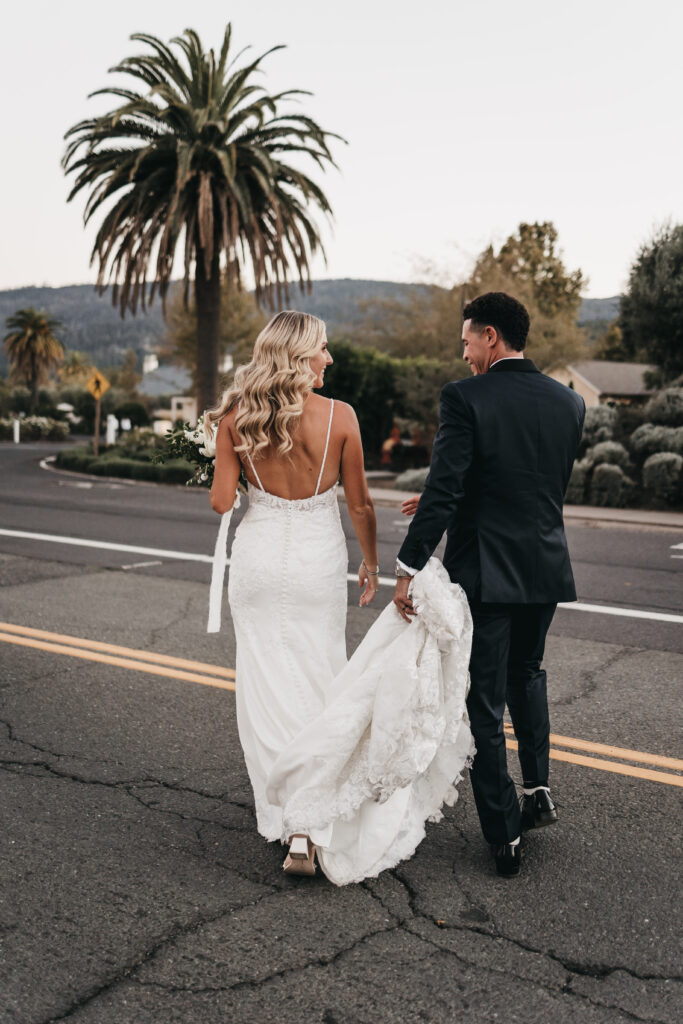 A bride in a white dress and a groom in a dark suit walk hand-in-hand down an empty street, with palm trees and hills in the background.