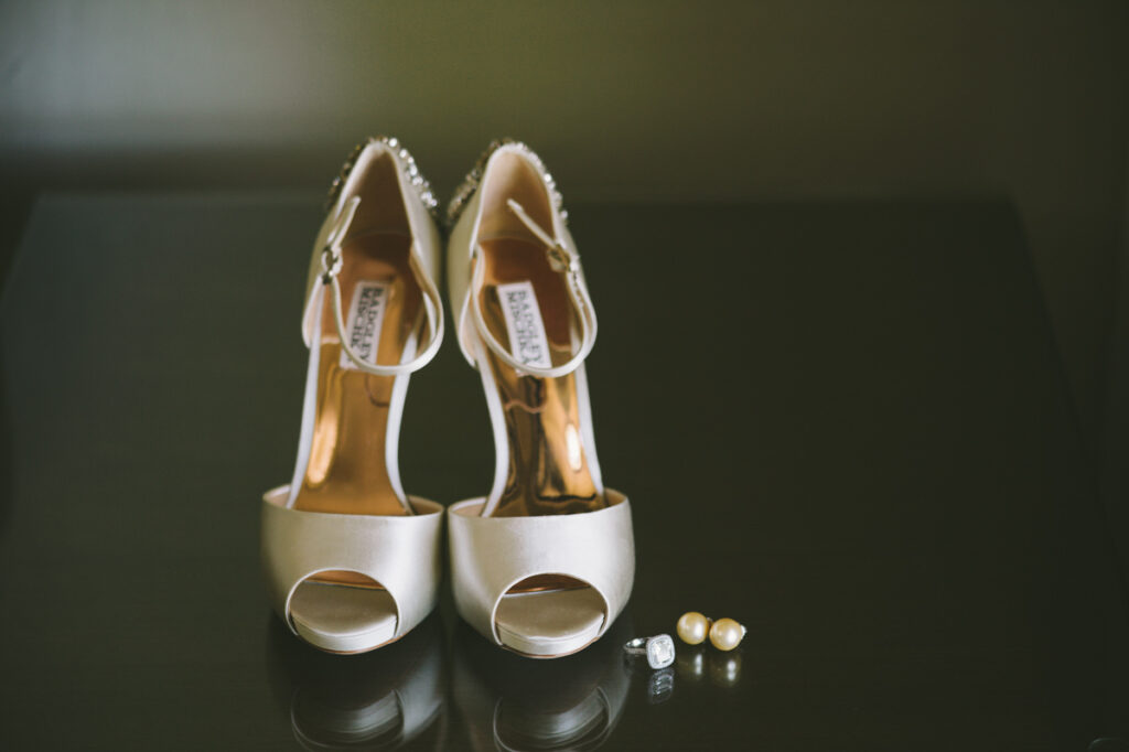 A pair of ivory high-heeled shoes is displayed on a dark surface with two rings and three pearl earrings arranged beside them.