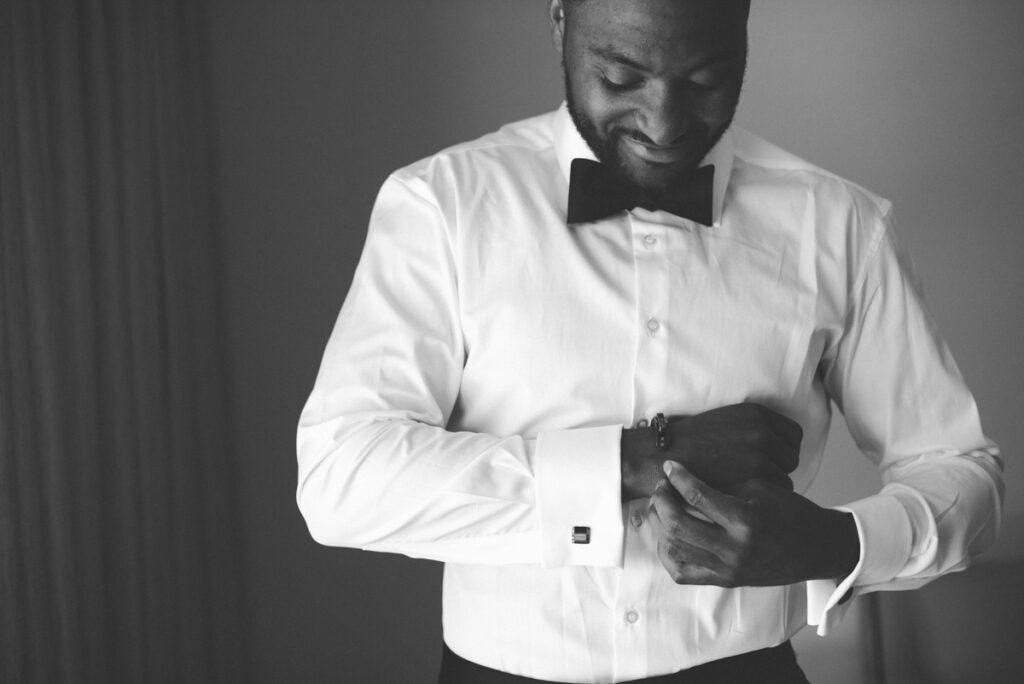 A man in a white dress shirt and black bow tie fastens his cufflink, smiling slightly. The photo is in black and white.