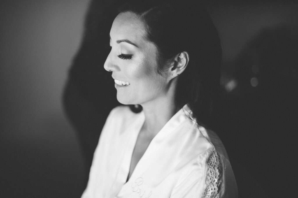A woman in a white robe with "Bride" embroidered on it smiles while looking down, photographed in black and white.