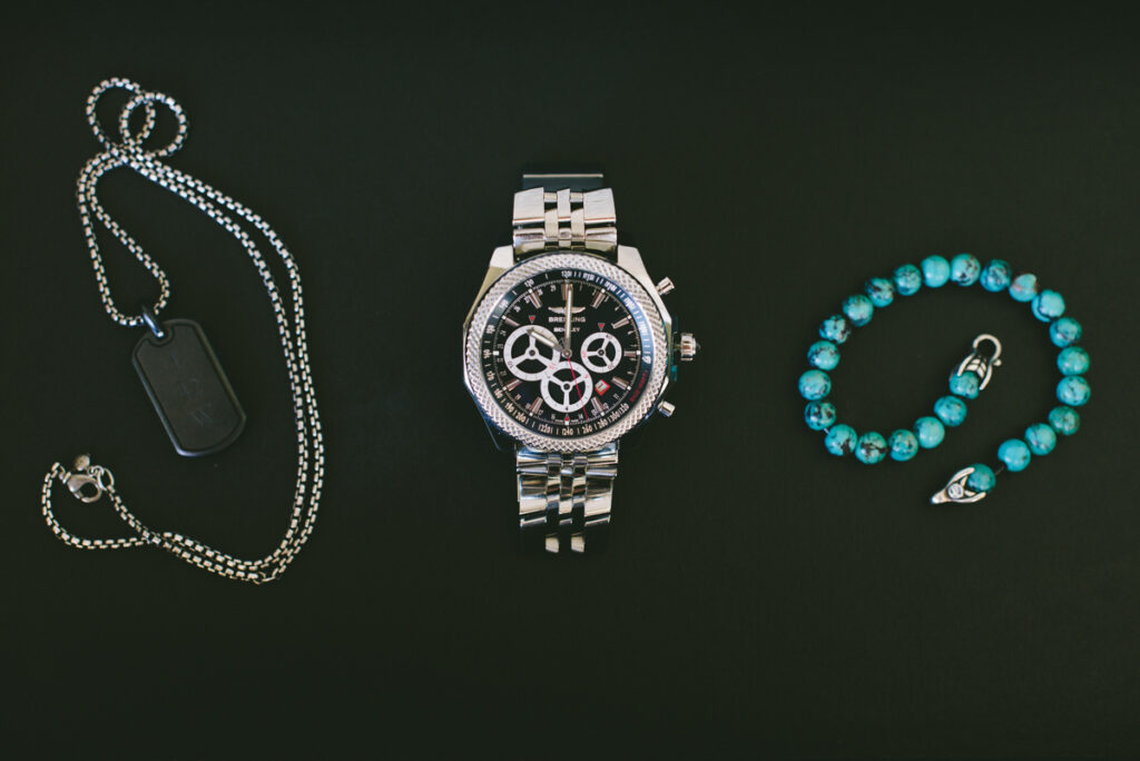 A silver wristwatch is centered between a silver dog tag necklace on the left and a turquoise bead bracelet on the right, all placed on a black background.