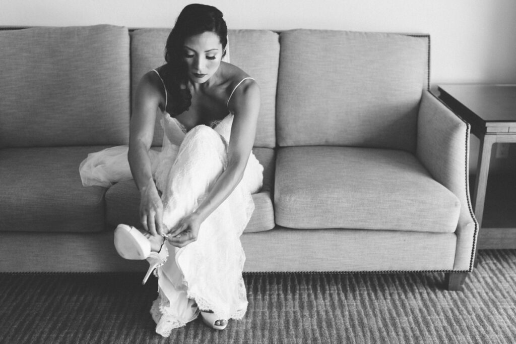 A woman in a wedding dress sits on a sofa and fastens her shoe. The photo is in black and white.