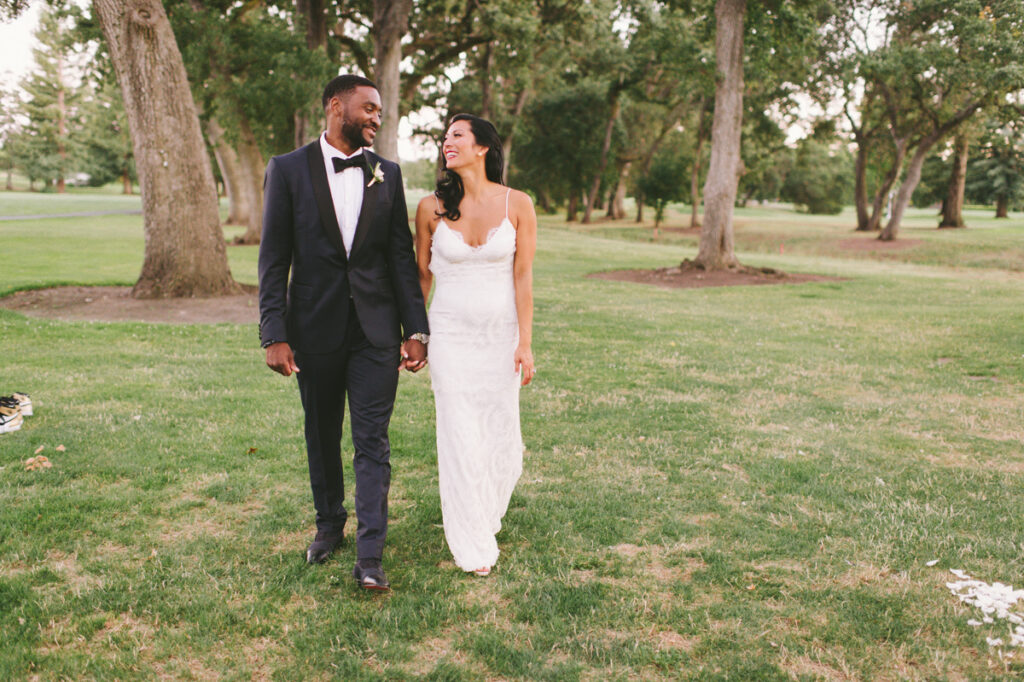 A bride in a white dress and a groom in a black suit walk hand in hand on grass in a park, smiling at each other.