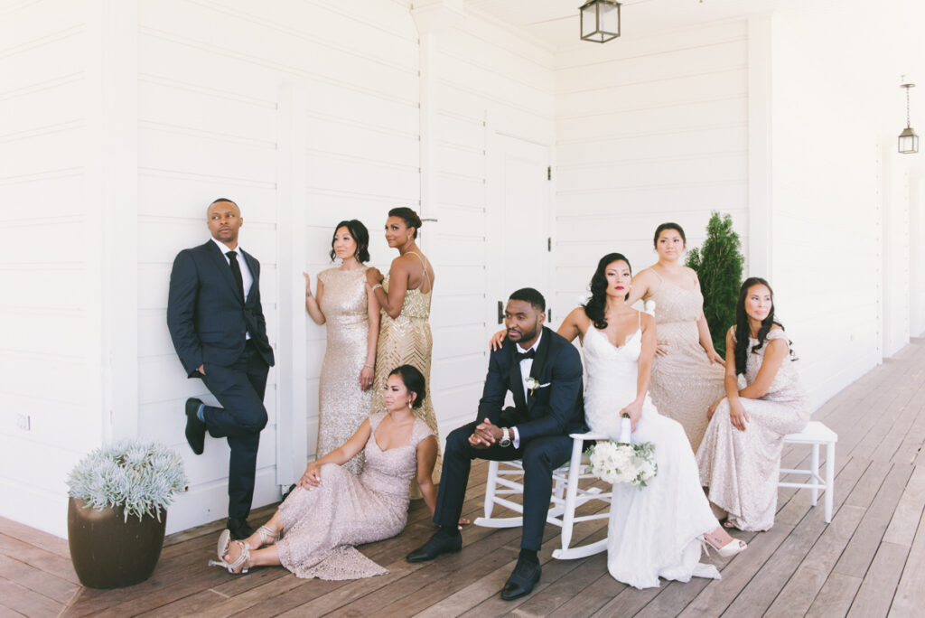 A wedding party poses on a wooden deck; the bride and groom sit at the center, surrounded by bridesmaids in light dresses and a groomsman in a navy suit against a white wall.