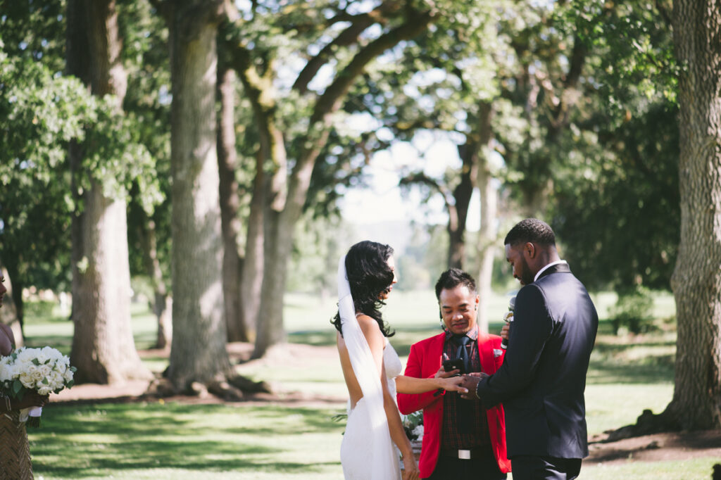 A couple stands with an officiant during an outdoor wedding ceremony in a wooded area, exchanging rings.