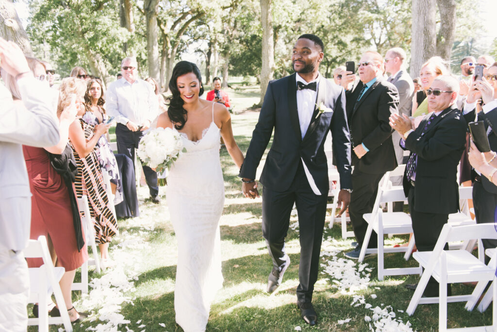 A bride and groom walk down an outdoor aisle lined with white chairs and flower petals, while guests stand, smile, and applaud.