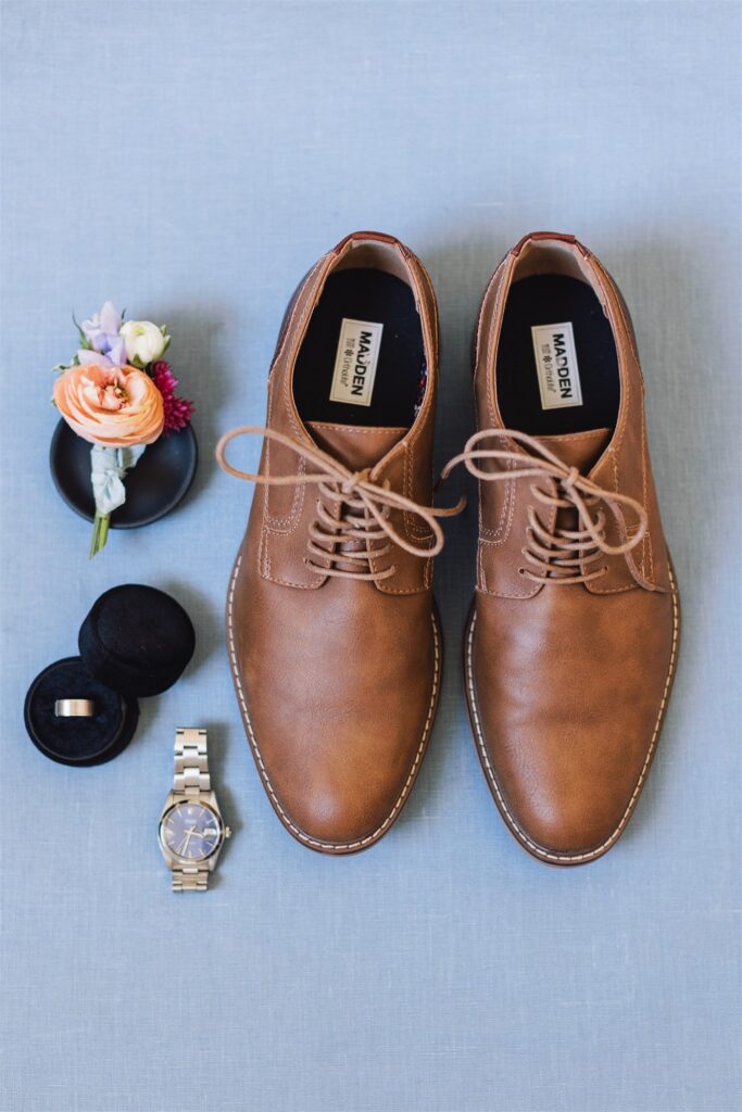 A pair of brown dress shoes, a boutonnière, a ring in a box, and a silver wristwatch arranged on a light blue surface.