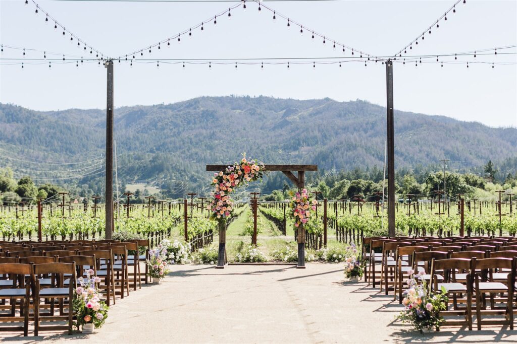 Outdoor wedding ceremony setup in a vineyard with rows of chairs, a floral arch, and string lights, set against a backdrop of green hills and clear sky.