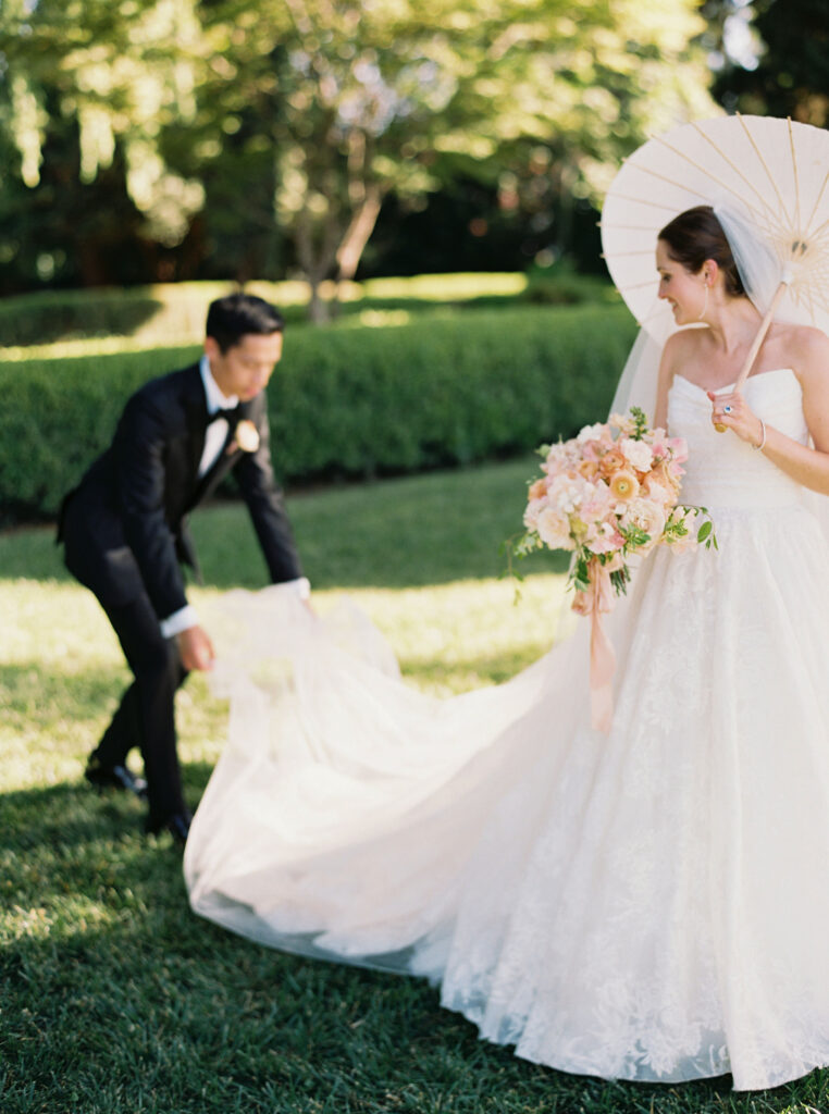 A bride holding a bouquet and a parasol stands on grass while a groom in a suit adjusts her long white dress train in a sunlit garden.