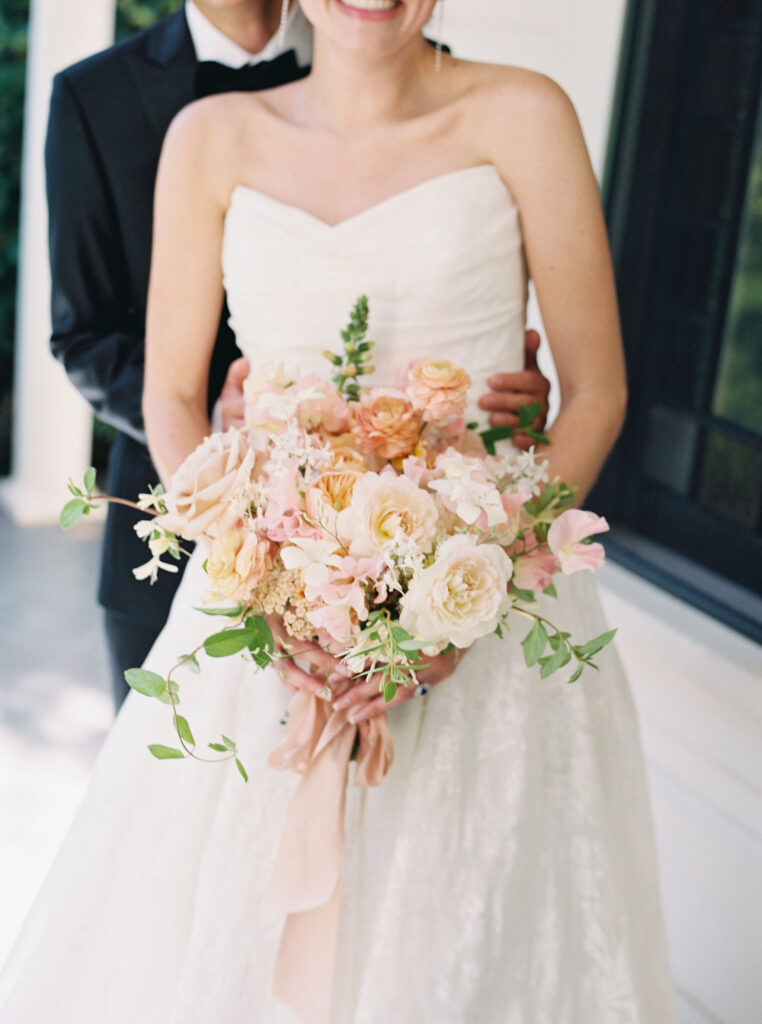 A bride in a strapless white dress holds a pastel bouquet, with a man in a black suit standing closely behind her.