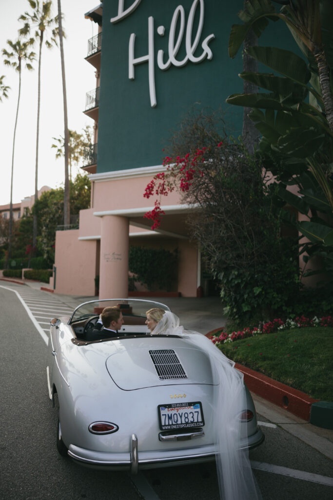 A bride and groom sit in a vintage silver convertible parked near a pink and green building with a "Beverly Hills" sign.