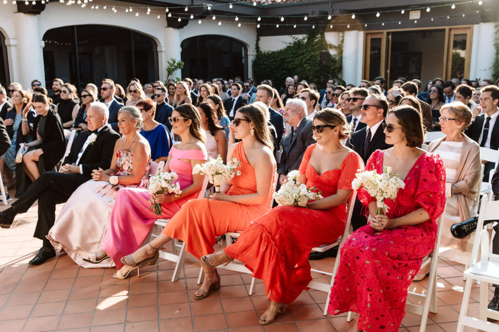 A seated wedding audience in formal attire, with several women in colorful dresses and sunglasses holding bouquets in the front row, outdoors under string lights.