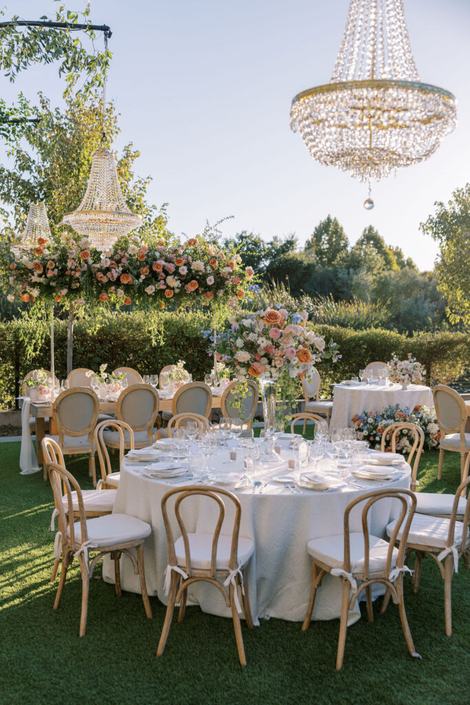 Outdoor wedding reception setup with round tables covered in white linens, elegant chairs, floral centerpieces, and hanging crystal chandeliers, surrounded by greenery.