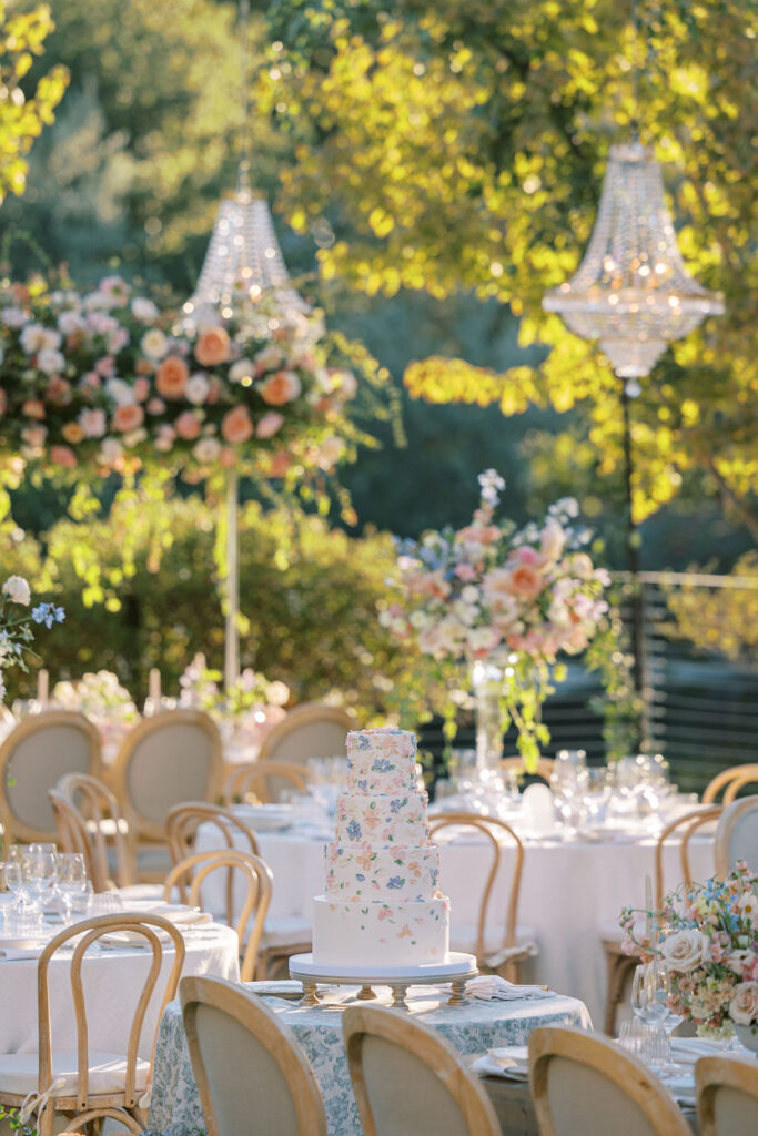 Outdoor wedding reception setup with round tables, floral centerpieces, chandeliers, and a tiered floral cake in the center, surrounded by chairs and greenery in bright sunlight.