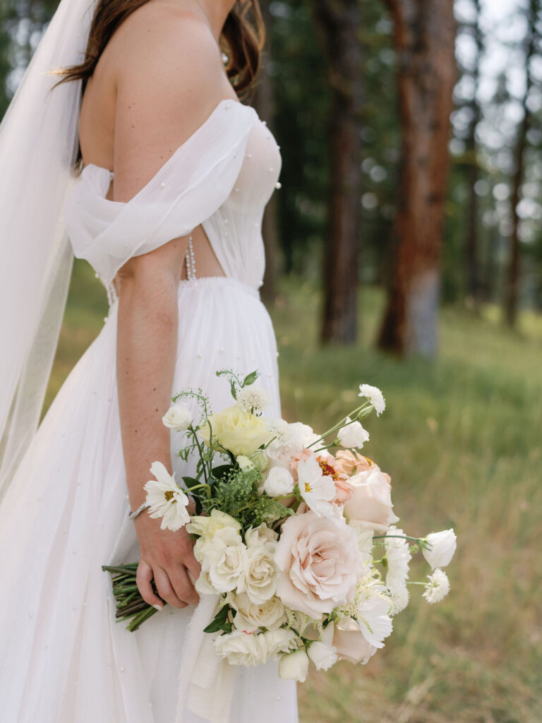 A bride in a white gown holds a bouquet of white and pale pink flowers while standing outdoors in a grassy, wooded area.
