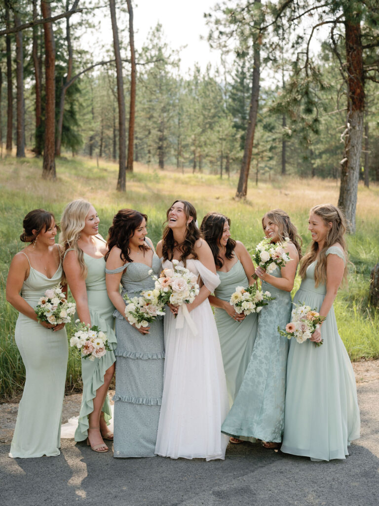 A bride in a white dress stands outdoors with five bridesmaids in light green dresses, all holding bouquets and smiling at each other. Trees and grass are visible in the background.