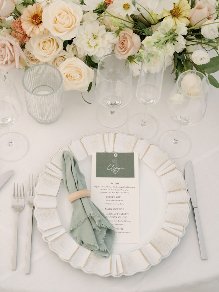 An elegant table setting with a white plate, a green cloth napkin in a ring, a printed menu, silver cutlery, glassware, and a floral centerpiece with white and blush flowers.