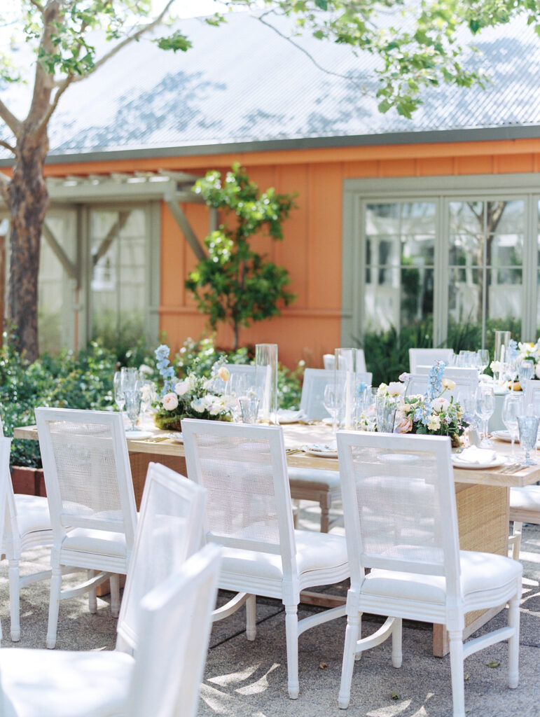 An outdoor dining table set with white chairs, glassware, and floral centerpieces, situated near an orange building with green trim and surrounded by trees and greenery.