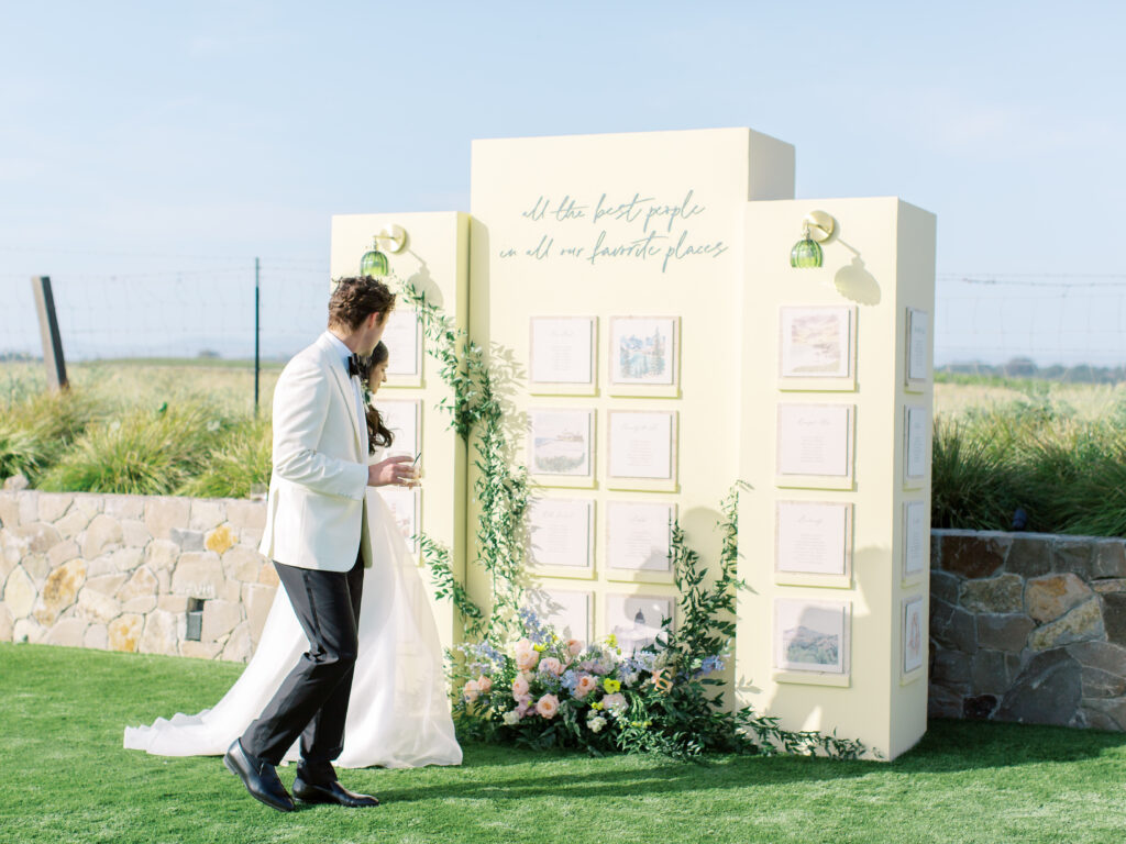 A couple in formal wedding attire stands beside a cream-colored display board with framed cards and greenery, outdoors on grass with a stone wall and blue sky in the background.