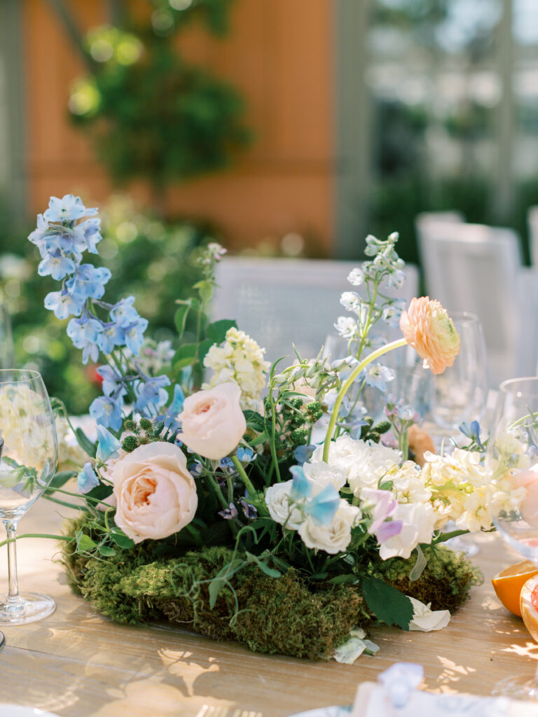A floral centerpiece with pink roses, blue delphiniums, and white flowers arranged on a moss base, set on a wooden table with glassware.