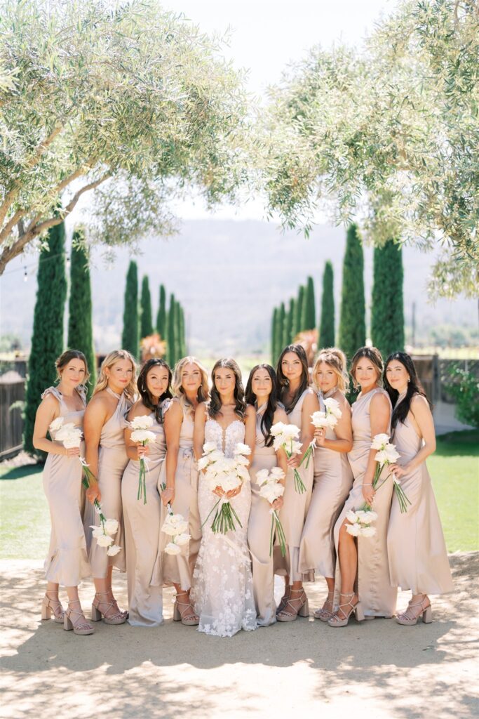 A bride in a white dress stands with ten bridesmaids in matching light-colored dresses, all holding flowers, posed outdoors amid trees and greenery.
