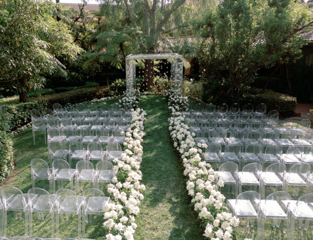 An outdoor wedding ceremony setup with rows of clear chairs and white flowers lining the grass aisle, leading to a floral arch under green trees.