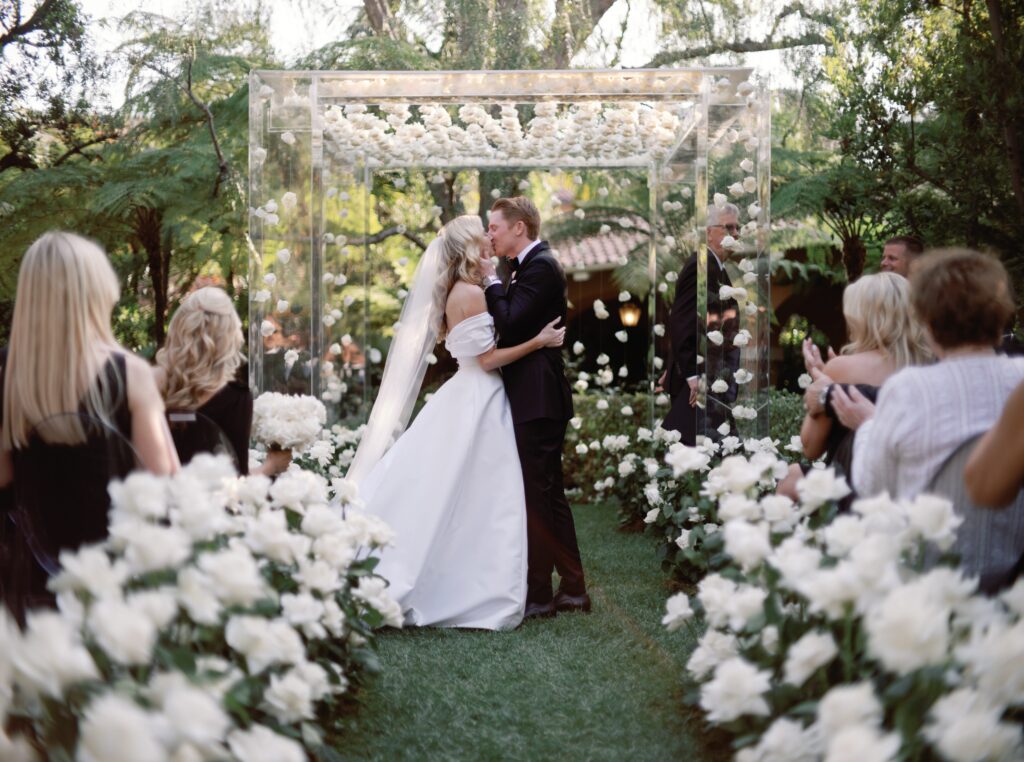 A bride and groom share a kiss at their outdoor wedding ceremony, surrounded by white flowers and applauding guests under a clear floral arch.