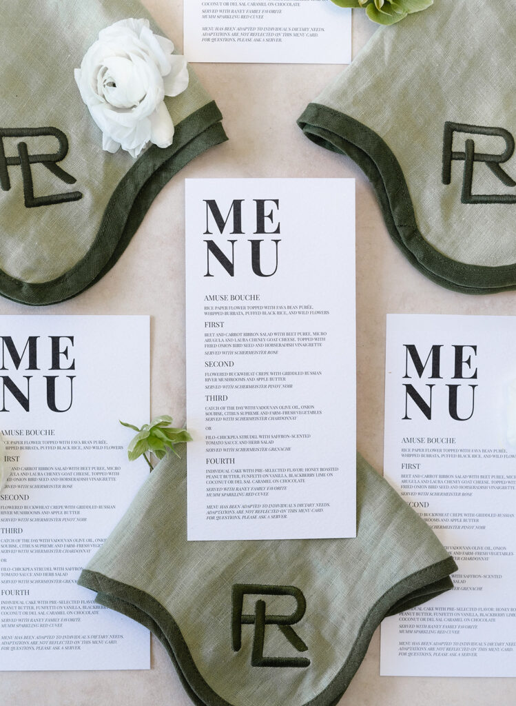 Overhead view of printed menus surrounded by green monogrammed napkins and white flowers on a light surface.