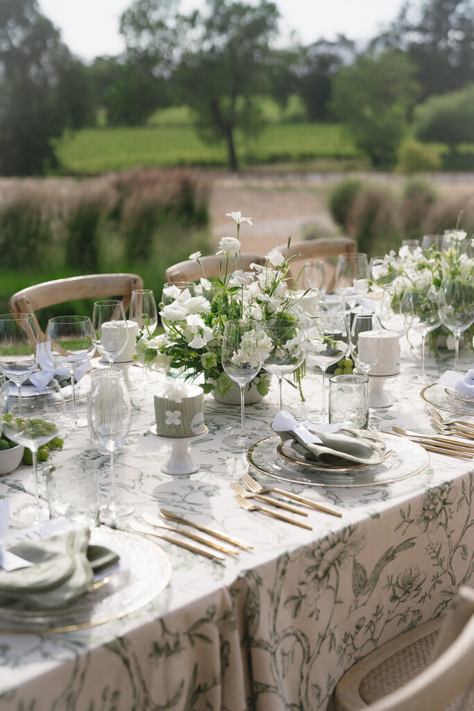 A formal outdoor dining table set with glassware, gold cutlery, floral centerpieces, and patterned tablecloth, with greenery and trees in the background.