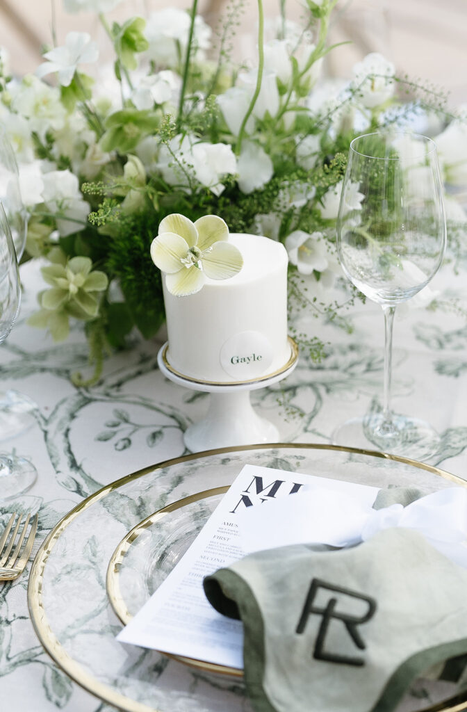 A small white cake with “Gayle” label sits on a stand beside green and white flowers, wine glasses, and a place setting with a menu and monogrammed napkin.