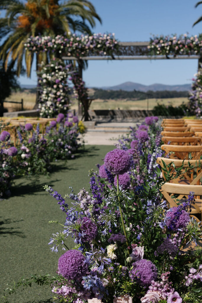 Outdoor wedding ceremony setup with rows of wooden chairs and an aisle decorated with purple flowers, under a floral arch, with palm trees and mountains in the background.
